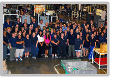 A group of floor employees on the shop floor. There is a sea of navy shirts with a pink tshirt and a white tshirt on the left.
